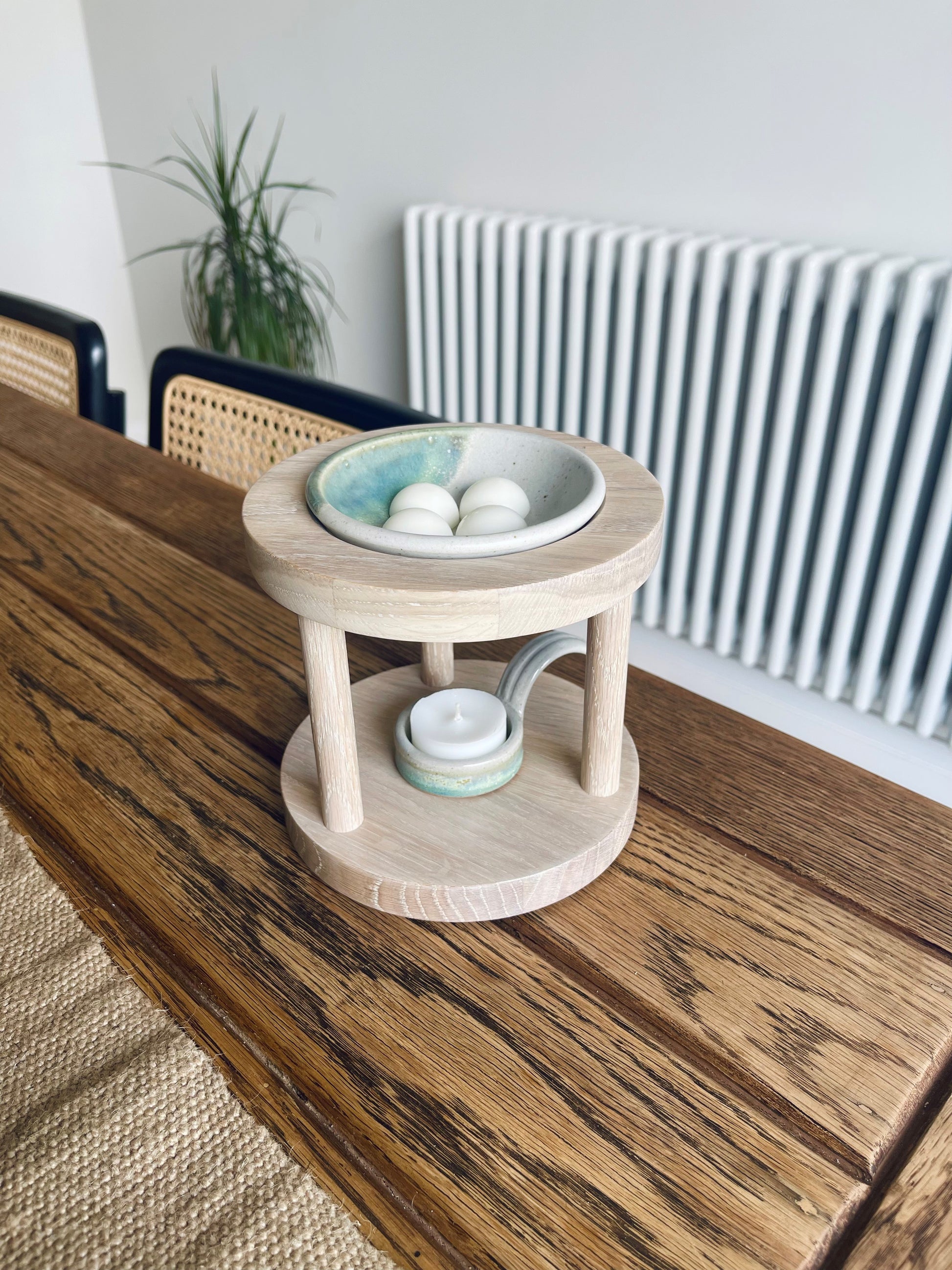 Round shaped wooden wax melt burner in clean limed oak shade with sage glazed pottery tealight spoon and wax melt dish on a windowsill