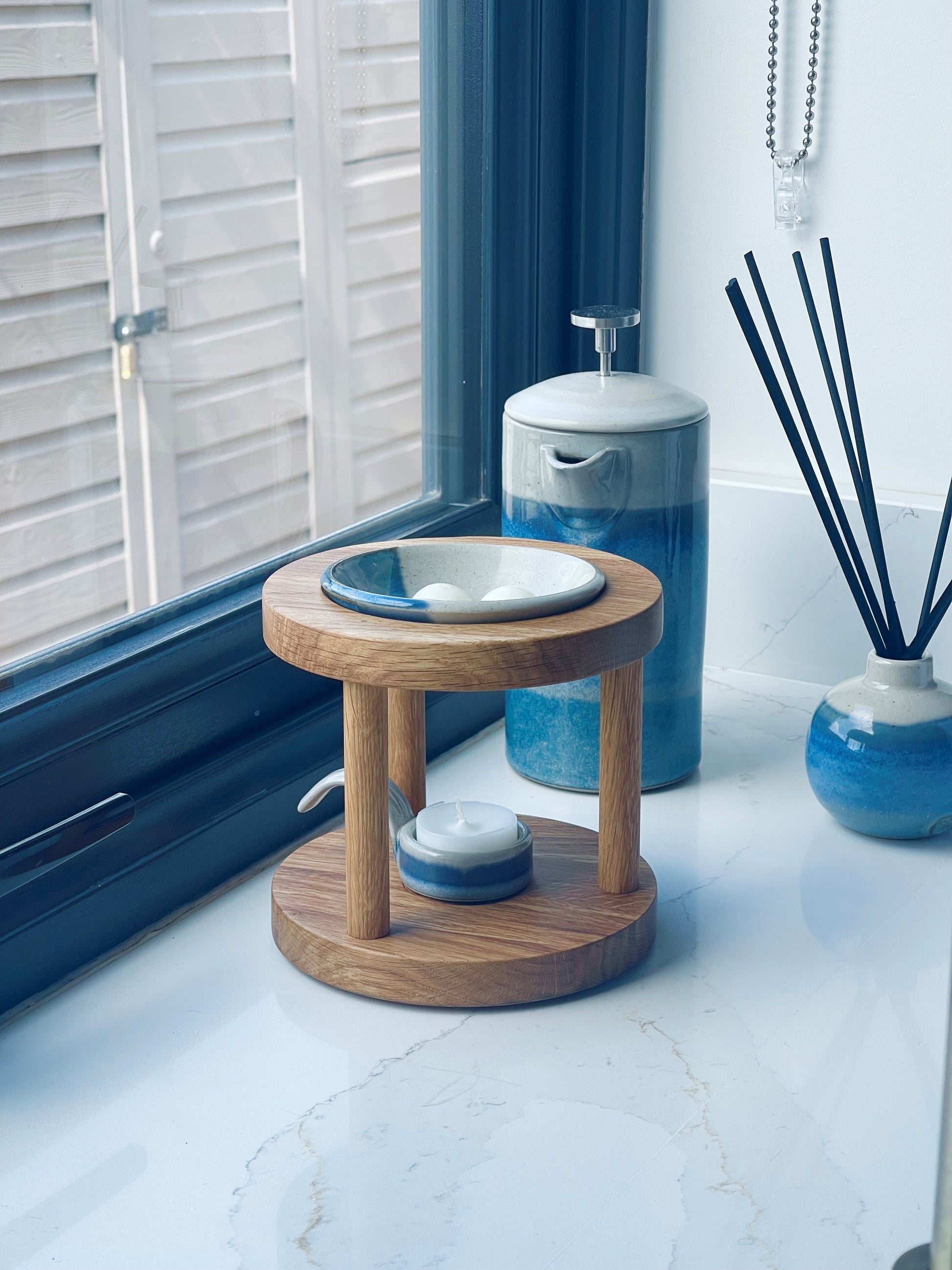 Round shaped wooden wax melt burner in warm oiled oak shade with blue glazed pottery tealight spoon and wax melt dish on a windowsill